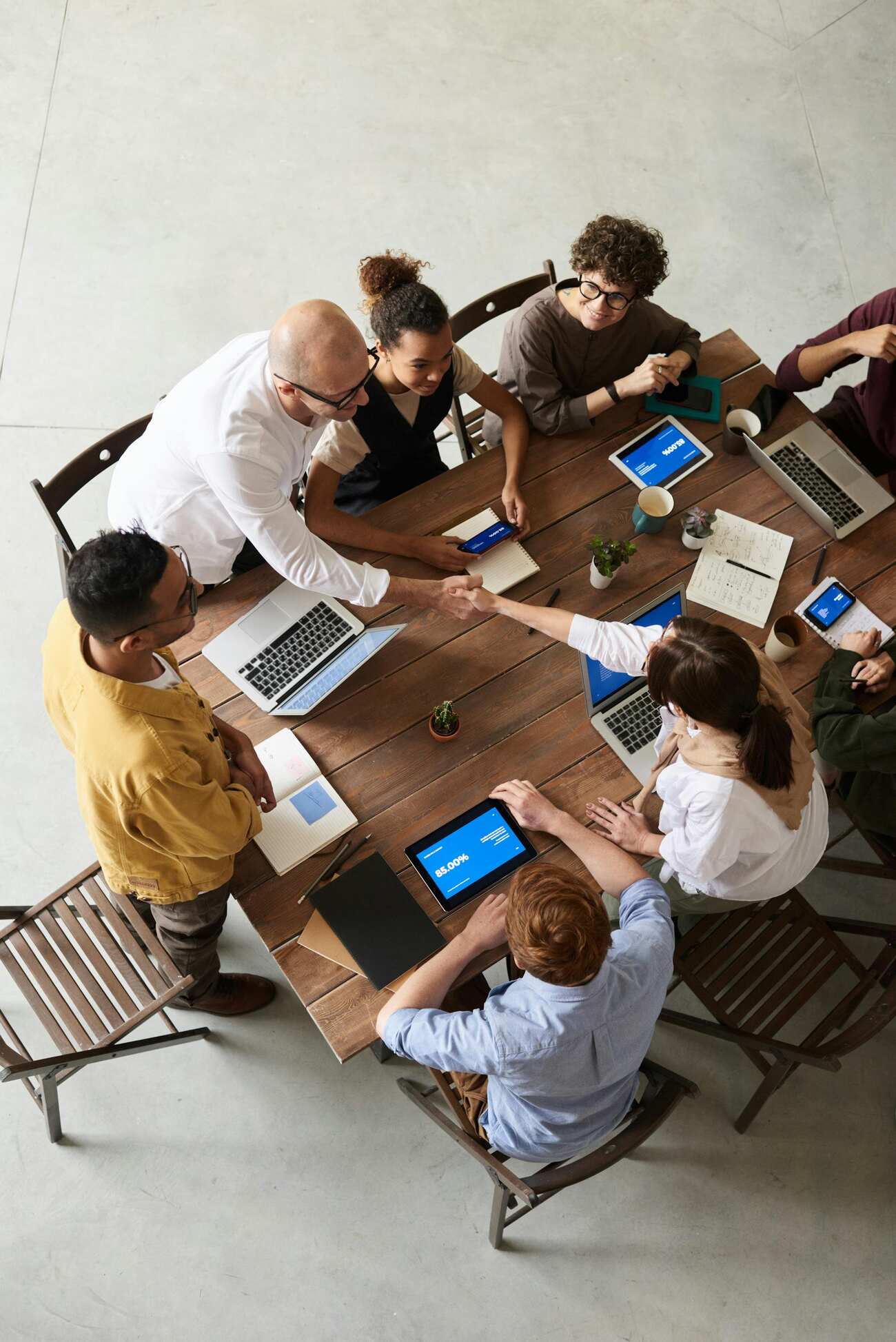 A diverse team gathered around a wooden table, shaking hands and collaborating on laptops and tablets