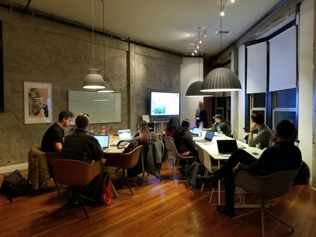 A business meeting in a modern, industrial-style room with a presenter leading a discussion on a large screen