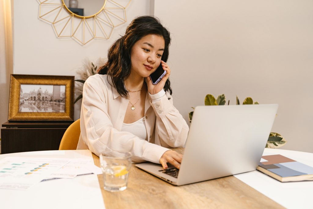 A woman in a beige blouse sits at a wooden desk, talking on her smartphone while working on a laptop