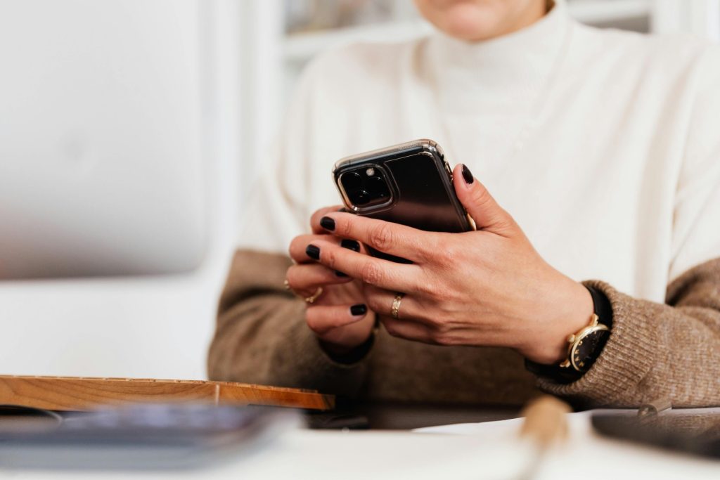 A close-up of a person's hands holding a smartphone. The person is wearing a beige sweater, black nail polish, and gold rings