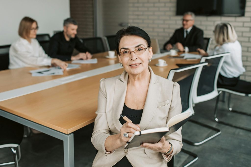 A confident older woman in a beige blazer sits at a conference table, holding a notebook and pen