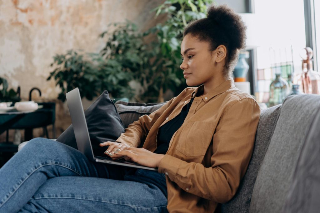 A woman in a brown jacket sitting on a couch, working on a laptop in a cozy, plant-filled room.