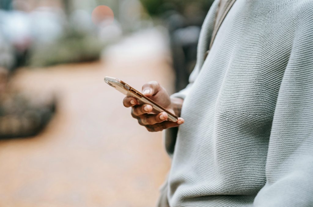 A person wearing a light-colored sweater is holding a smartphone while standing outdoors. The background is blurred, showing a paved walkway with trees.