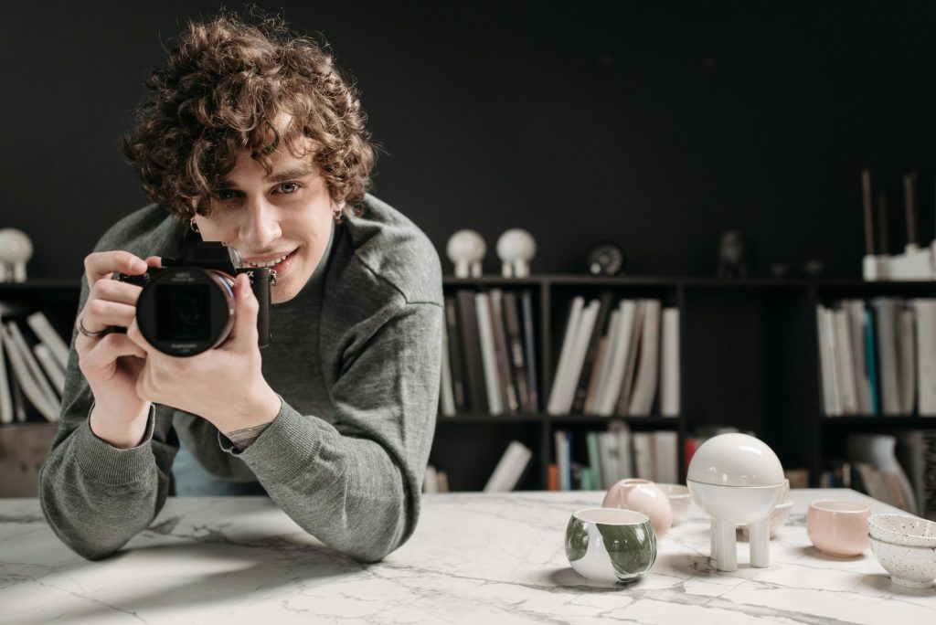 A young man with curly hair and a smile holds a professional camera, looking through the lens