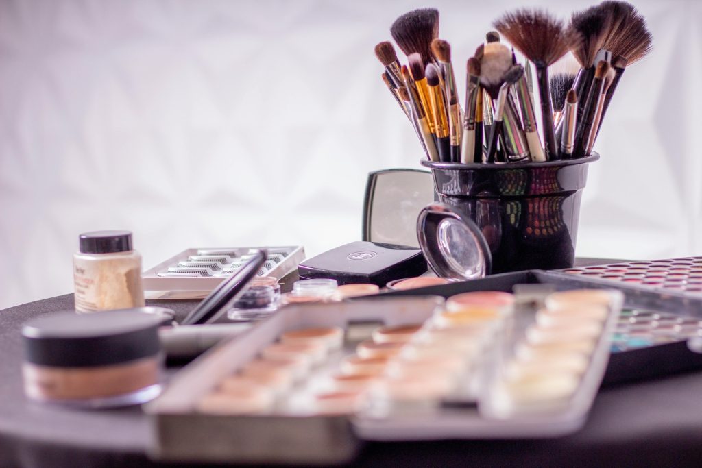 A collection of makeup products and tools displayed on a table, including brushes in a black container, eyeshadow palettes, foundation, and false eyelashes