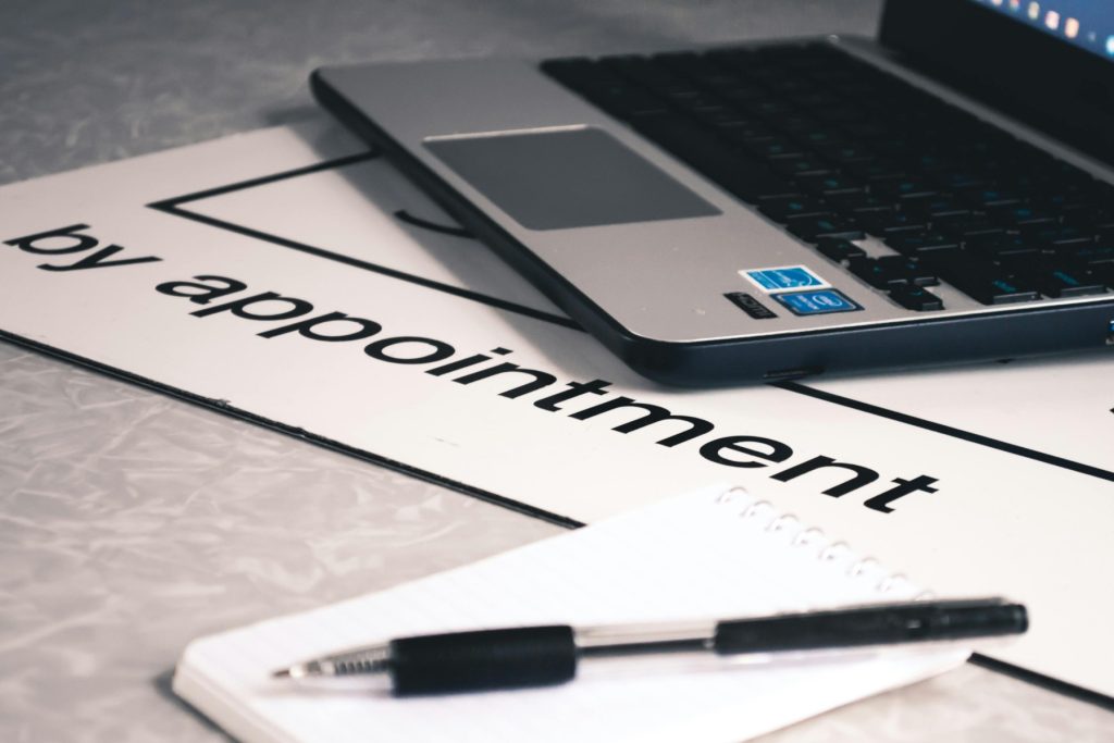 A laptop placed on a table with a sign that reads "by appointment," accompanied by a notebook and a pen, symbolizing appointment scheduling