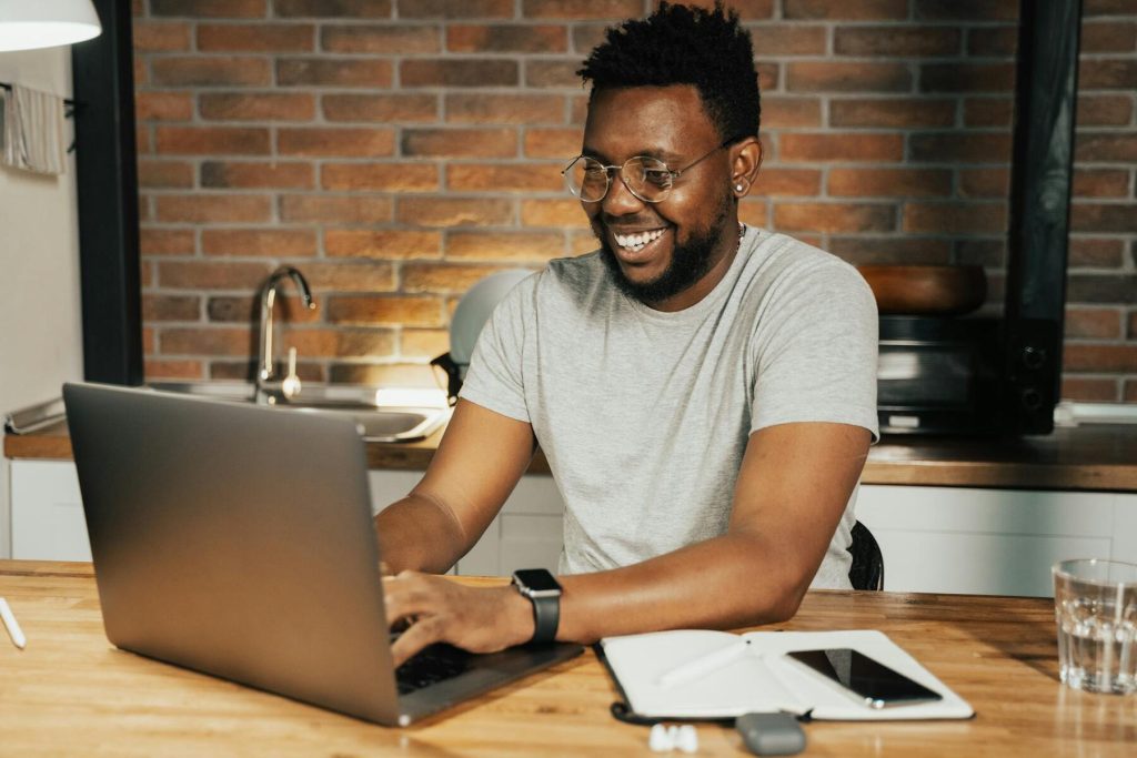 A smiling man sits at a table, working on a laptop