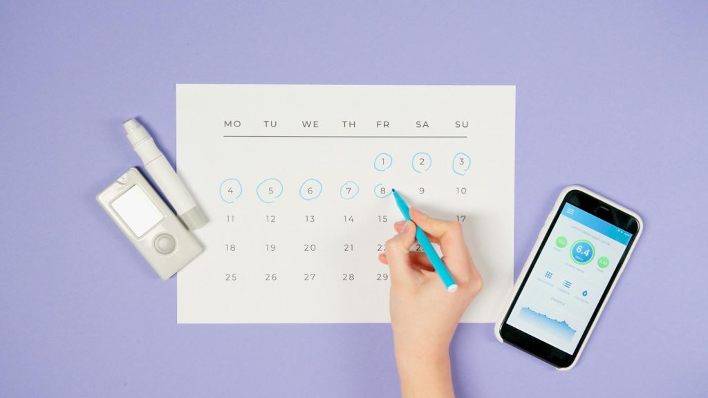A woman's hand writes on a calendar with a pen, while a smartphone rests nearby on a purple background